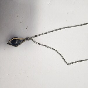 May include: A silver-toned necklace featuring a geometric pendant with a dark, spherical bead. The pendant is diamond-shaped with a thin frame. The chain is a delicate curb link style, suitable for everyday wear.