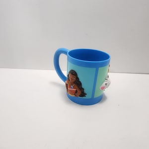 May include: A blue plastic mug featuring a cartoon character design. The mug has a blue handle and a light blue background with a cartoon character on the front. The mug is designed for children.