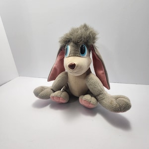 Puede incluir: Un peluche de conejo con pelaje gris, ojos azules grandes y orejas internas rosas. El conejo tiene una nariz marrón claro y patas con almohadillas rosas. Está sentado sobre una superficie blanca.