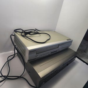 Może przedstawiać: Dwa urządzenia elektroniczne ułożone jedno na drugim. Odtwarzacz combo VCR/DVD Sylvania ze srebrnym przodem i beżowym korpusem znajduje się na czarnym odtwarzaczu DVD. Czarny przew&oacute;d zasilający jest przerzucony przez g&oacute;rę.