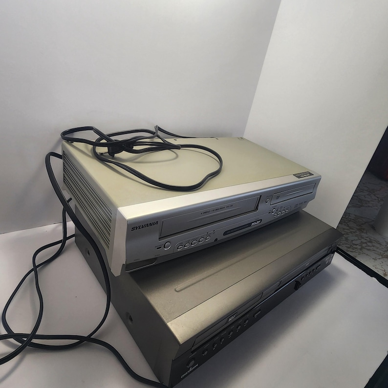 Może przedstawiać: Dwa urządzenia elektroniczne ułożone jedno na drugim. Odtwarzacz combo VCR/DVD Sylvania ze srebrnym przodem i beżowym korpusem znajduje się na czarnym odtwarzaczu DVD. Czarny przew&oacute;d zasilający jest przerzucony przez g&oacute;rę.