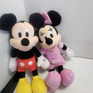 May include: A plush Mickey Mouse and Minnie Mouse toy set. Mickey is wearing red shorts with white polka dots and yellow shoes. Minnie is wearing a pink dress with white polka dots and pink shoes.