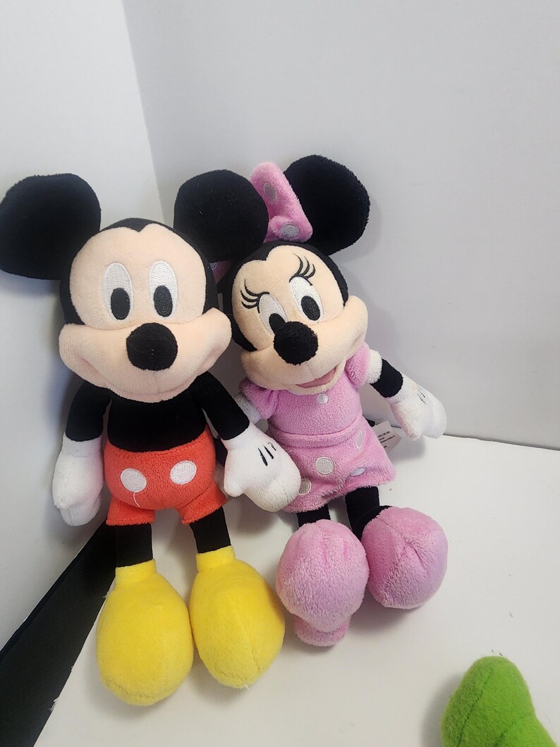 May include: A plush Mickey Mouse and Minnie Mouse toy set. Mickey is wearing red shorts with white polka dots and yellow shoes. Minnie is wearing a pink dress with white polka dots and pink shoes.