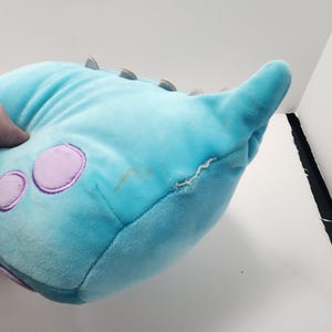 May include: A light blue monster-shaped plush toy with purple circular spots and small brown spikes. The toy has a pointed horn and a small tear on the side. The plush is made of a soft material.