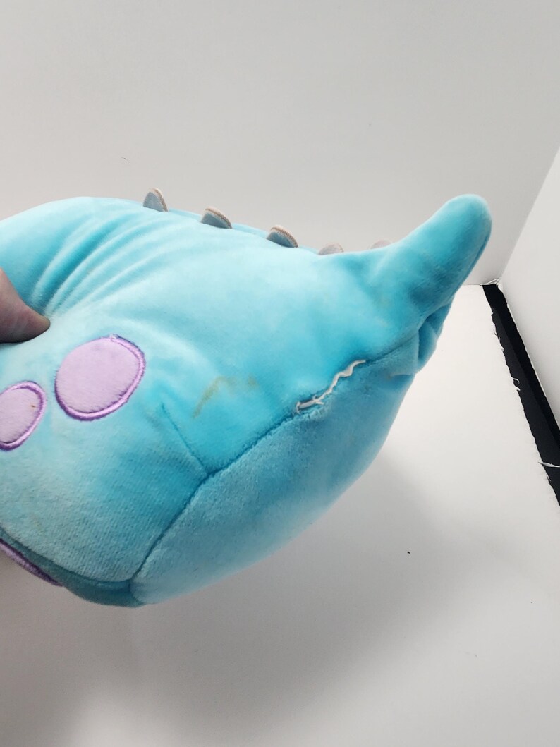 May include: A light blue monster-shaped plush toy with purple circular spots and small brown spikes. The toy has a pointed horn and a small tear on the side. The plush is made of a soft material.