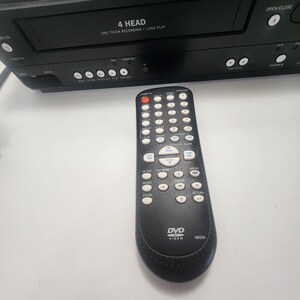 K&ouml;nnte beinhalten: Schwarzer Videorecorder mit einer 4-Kopf-One-Touch-Aufnahmefunktion und einer schwarzen Fernbedienung mit dem Text "DVD Video NB093" darauf.