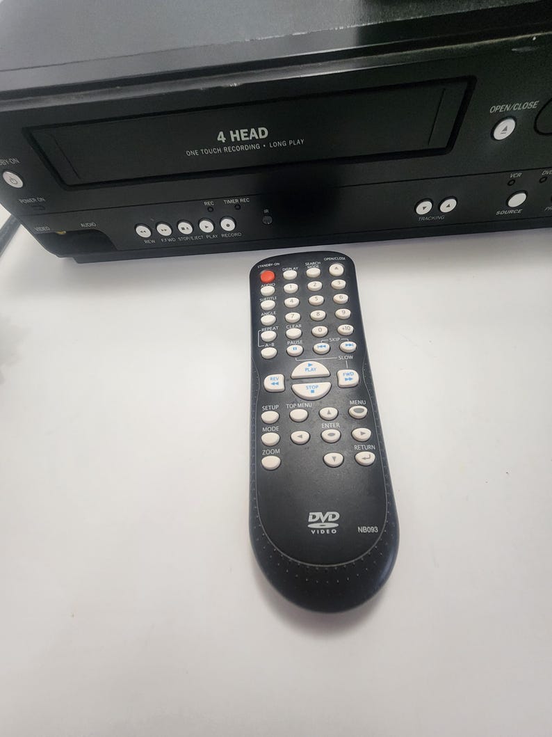 K&ouml;nnte beinhalten: Schwarzer Videorecorder mit einer 4-Kopf-One-Touch-Aufnahmefunktion und einer schwarzen Fernbedienung mit dem Text "DVD Video NB093" darauf.
