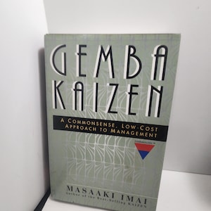 Puede incluir: Un libro titulado "GEMBA KAIZEN" con una cubierta verde claro. El título está en letras blancas grandes y en negrita. El subtítulo del libro dice "A Commonsense, Low-Cost Approach to Management." El autor es Masaaki Imai.