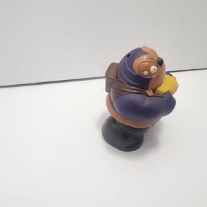 May include: A colorful figurine of a cartoon character with a brown head, purple body, and green base. The character wears a brown backpack and holds a yellow object. The figurine is set against a white background.