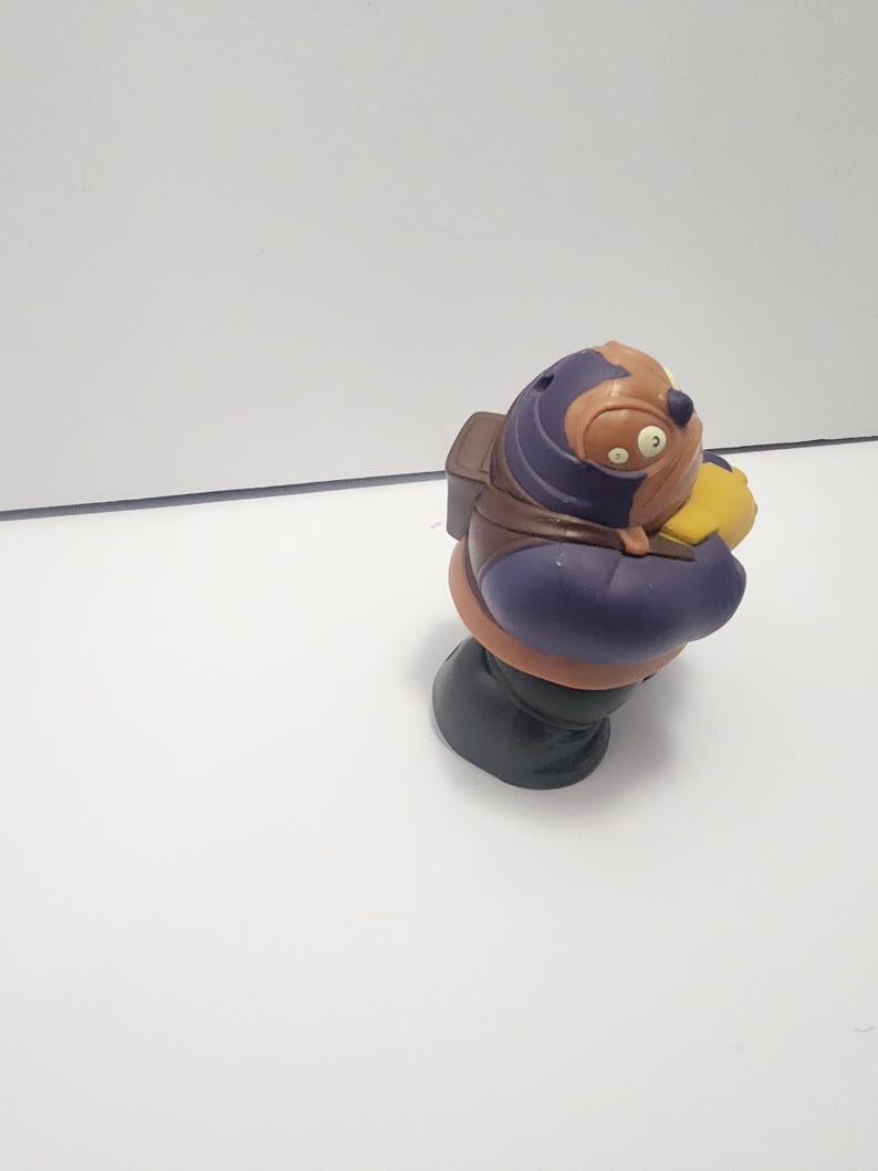 May include: A colorful figurine of a cartoon character with a brown head, purple body, and green base. The character wears a brown backpack and holds a yellow object. The figurine is set against a white background.