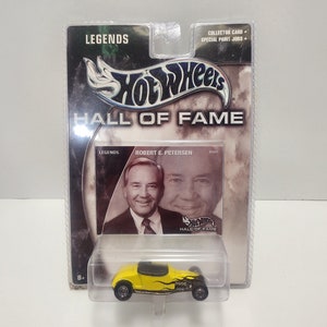 May include: A yellow Hot Wheels car with black flames on the side. The car is in a plastic package with a card featuring a photo of Robert E. Petersen, a legend in the car industry, and the text "Hot Wheels Hall of Fame".