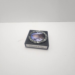May include: A silver-toned, circular decorative piece with an image of a building and a decorative border, set on a black rectangular base. The base has the text "Delivered As One".