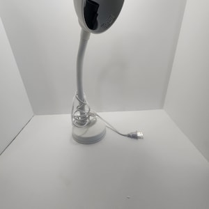 Peut inclure: Une lampe de bureau blanche avec un col de cygne flexible et un abat-jour rond blanc. La lampe a un cordon blanc et une prise blanche.