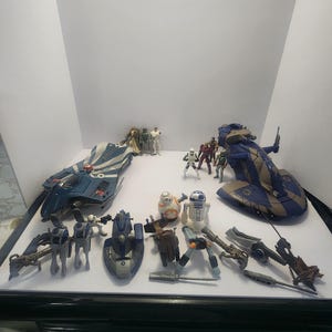 May include: A selection of Star Wars toys, featuring a blue and white starfighter, a large blue and brown transport, and several character figures such as BB-8 and R2-D2. The toys are displayed on a white surface.