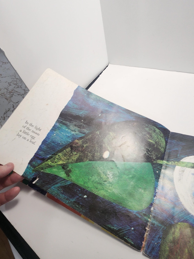 Puede incluir: Un libro abierto en una p&aacute;gina con una ilustraci&oacute;n colorida de una hoja verde, azul y amarilla con un huevo blanco en la parte superior. El texto en la p&aacute;gina dice "In the light of the moon a little egg lay on a leaf."