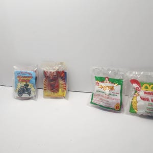 May include: Four sealed plastic bags containing children's toys. The bags feature colourful graphics and text, including "Masked Rider" with a motorcycle, a red figure, and "Spider-Man" with a green background. The toys appear to be from a fast-food promotion.