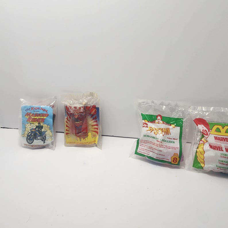 May include: Four sealed plastic bags containing children's toys. The bags feature colourful graphics and text, including "Masked Rider" with a motorcycle, a red figure, and "Spider-Man" with a green background. The toys appear to be from a fast-food promotion.