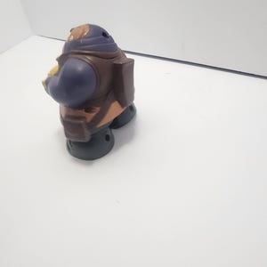 May include: A small, plastic toy figure in a seated position. The figure has a purple head and arms, a brown torso, and black feet. It appears to be a character from a children's show or movie.