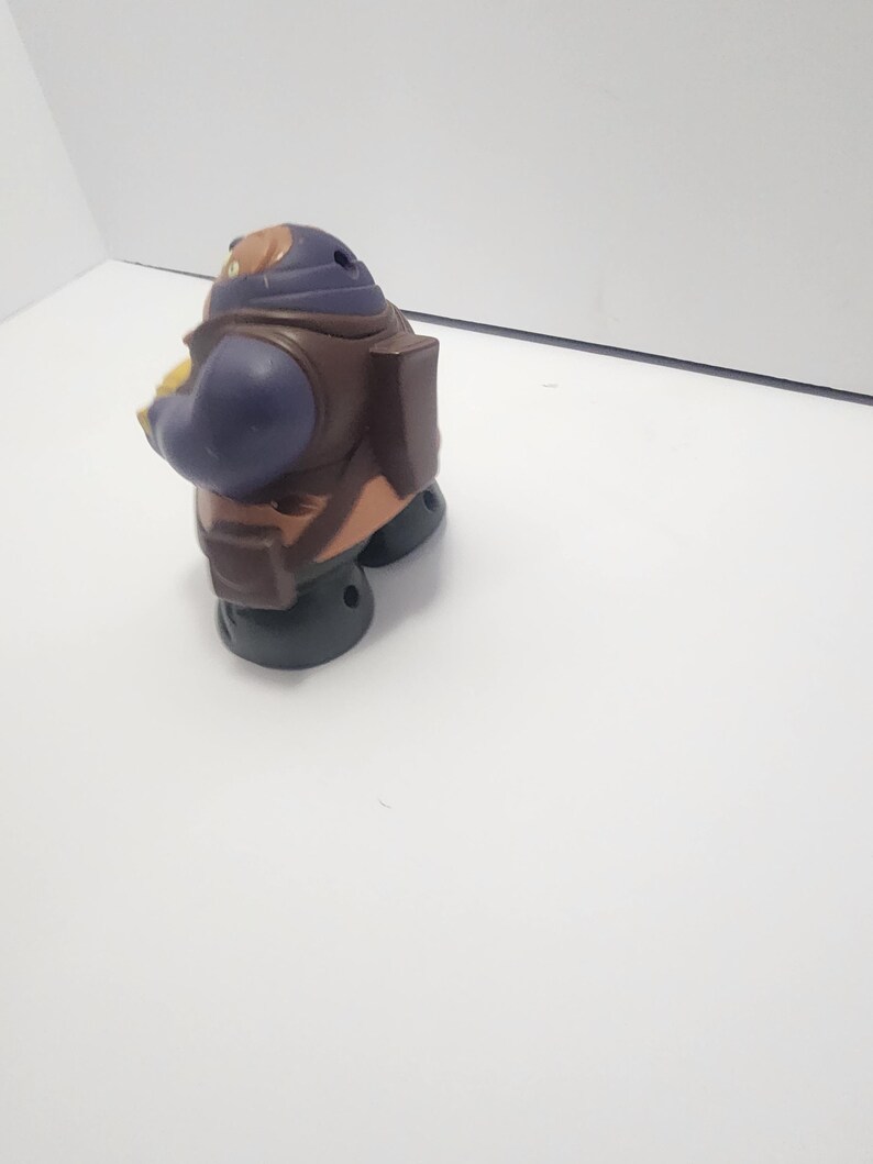 May include: A small, plastic toy figure in a seated position. The figure has a purple head and arms, a brown torso, and black feet. It appears to be a character from a children's show or movie.