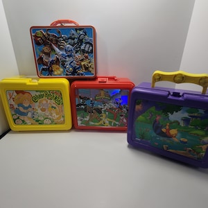 May include: Four vintage lunchboxes featuring various cartoon characters. The lunchboxes are yellow, red, purple, and orange. The yellow lunchbox has a Cabbage Patch Kids design, the red lunchbox has a Power Rangers design, the purple lunchbox has a Winnie the Pooh design, and the orange lunchbox has a cartoon character design.