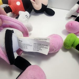May include: A set of four plush Disney Mickey Mouse characters. The characters are pink, green, black and red. The characters are all in different poses. The tag on the pink character reads "Surface Washable Only Air Dry. Contents: All New Materials. Polyester Fibres, Polyethylene Fibres & Polyester Felt. Reg. No. PA-15431/400. Made in China."