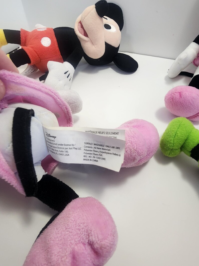 May include: A set of four plush Disney Mickey Mouse characters. The characters are pink, green, black and red. The characters are all in different poses. The tag on the pink character reads "Surface Washable Only Air Dry. Contents: All New Materials. Polyester Fibres, Polyethylene Fibres & Polyester Felt. Reg. No. PA-15431/400. Made in China."