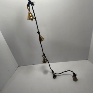 May include: A decorative hanging ornament featuring small, gold-colored bells strung along a black cord. The bells are of varying sizes and shapes, with a shiny, metallic finish. The cord is knotted between the bells.