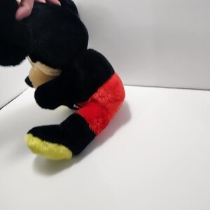 May include: A plush toy of a cartoon mouse, featuring a black body, red shorts, and yellow shoes. The toy's head is black with tan ears and a tan face. The toy is partially visible, with the hand of a person holding it.