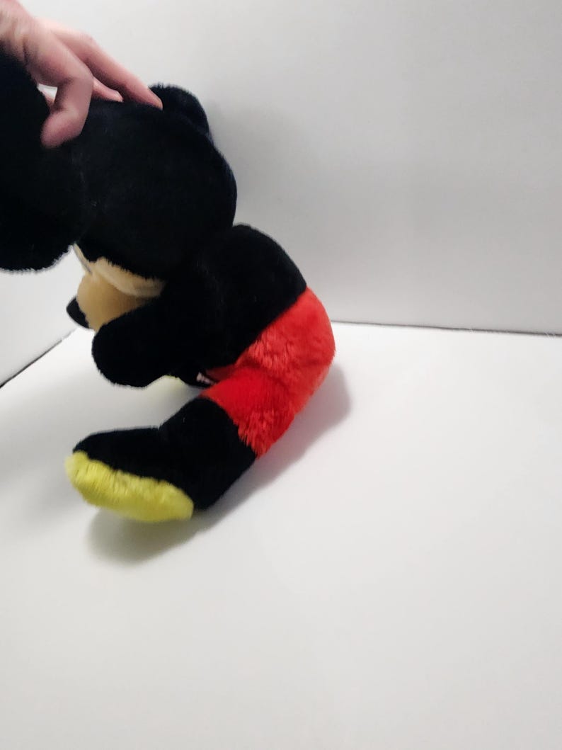 May include: A plush toy of a cartoon mouse, featuring a black body, red shorts, and yellow shoes. The toy's head is black with tan ears and a tan face. The toy is partially visible, with the hand of a person holding it.