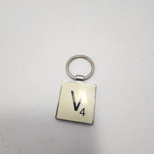 May include: A square keychain with a silver-colored ring and frame. The keychain features a cream-colored tile with the letter "V" and the number "4" in black. The tile is set within a silver-colored metal frame.
