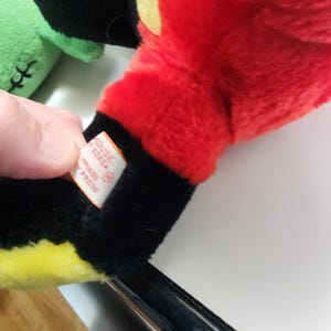 May include: Close-up of a plush toy's red hand with yellow and black accents. The hand is part of a larger toy, with a tag visible. The toy is on a white surface.