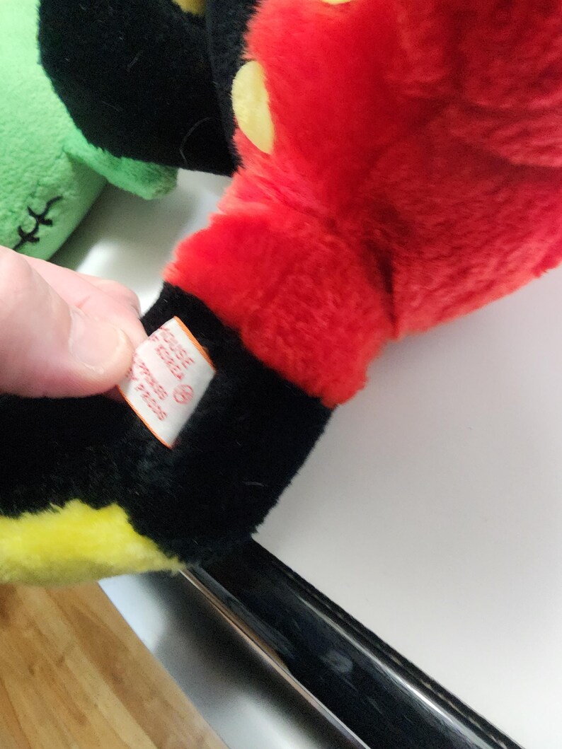 May include: Close-up of a plush toy's red hand with yellow and black accents. The hand is part of a larger toy, with a tag visible. The toy is on a white surface.