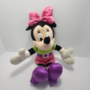 May include: A plush Minnie Mouse doll with a black head, large pink bow, and white gloves. She wears a green shirt, pink skirt, and purple shoes. The doll has a black nose and large, round eyes.