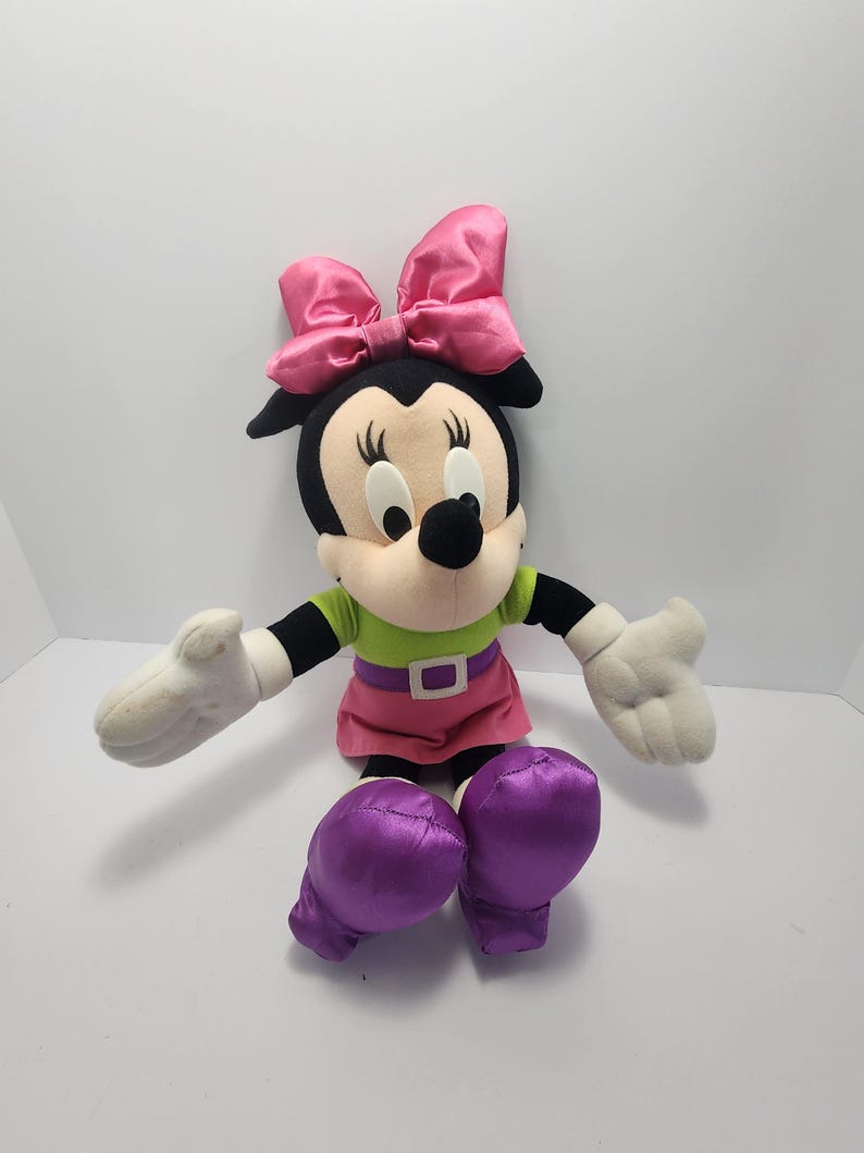 May include: A plush Minnie Mouse doll with a black head, large pink bow, and white gloves. She wears a green shirt, pink skirt, and purple shoes. The doll has a black nose and large, round eyes.