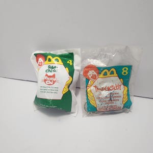 May include: Two sealed McDonald's Happy Meal toys. One is a green package with a cat graphic and the text "Meow Chi". The other is a clear package with a Tamagotchi keychain. Both packages have the McDonald's logo.
