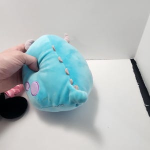 May include: A light blue plush monster toy with purple spots and small horns. It features a row of small, light-coloured spikes along its back. The toy is held by a hand.
