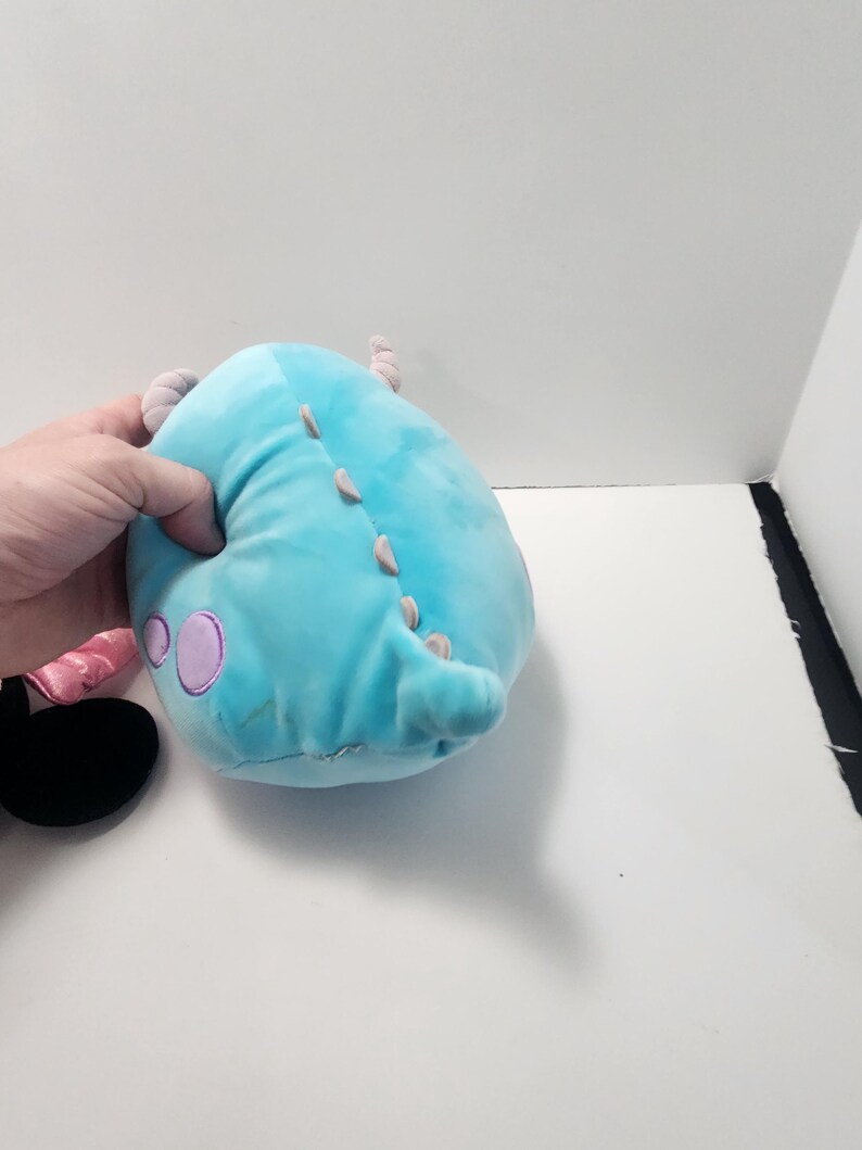 May include: A light blue plush monster toy with purple spots and small horns. It features a row of small, light-coloured spikes along its back. The toy is held by a hand.