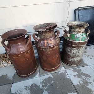 Puede incluir: Tres lecheras metálicas antiguas con asas. Las lecheras tienen un aspecto desgastado y oxidado, con diferentes tonos de pintura marrón y verde. Las lecheras son cilíndricas con una parte superior ensanchada y están dispuestas sobre una superficie de baldosas grises.