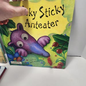 Puede incluir: Libro infantil titulado "Lucky Sticky Anteater" con una ilustraci&oacute;n de oso hormiguero de dibujos animados. La portada presenta un fondo amarillo con detalles verdes, un oso hormiguero morado y bayas rojas.