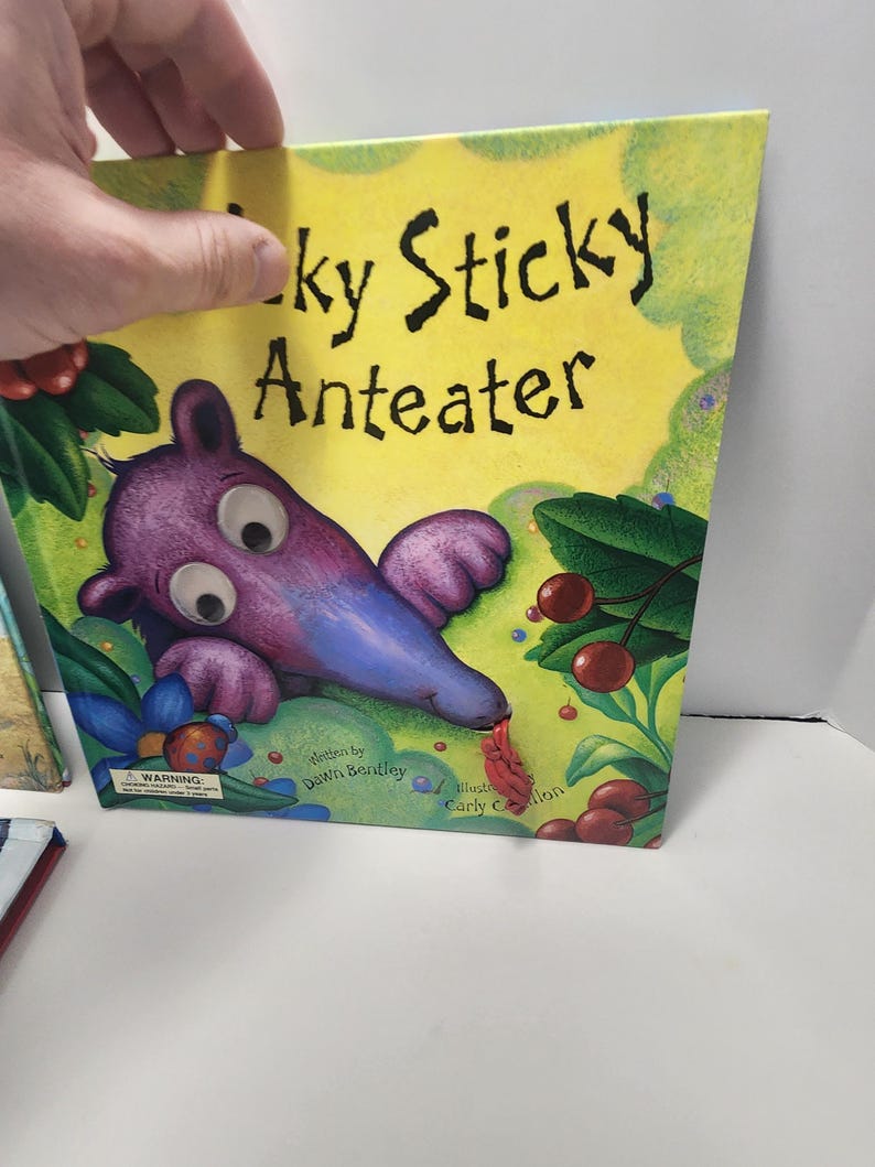 Puede incluir: Libro infantil titulado "Lucky Sticky Anteater" con una ilustraci&oacute;n de oso hormiguero de dibujos animados. La portada presenta un fondo amarillo con detalles verdes, un oso hormiguero morado y bayas rojas.
