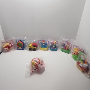 Puede incluir: Una colección de nueve juguetes de plástico coloridos en envases individuales de plástico transparente. Los juguetes están basados en un programa de televisión infantil y presentan personajes del programa. Los juguetes están en varias poses e incluyen un camión amarillo, un personaje azul, un personaje rosa y un personaje verde.