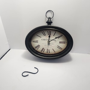 May include: An oval-shaped black wall clock with Roman numerals and the words "CHEVAL NOIR" on the face. The clock has a loop at the top for hanging. A small, black, curved hook sits below the clock.