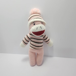 May include: A handmade sock monkey doll wearing a pink and brown striped sweater and matching hat with a pink pom-pom. The doll has a white face, white hands, and a red mouth. The doll is standing upright.