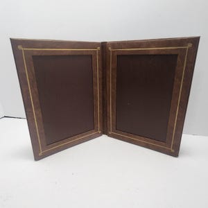 Peut inclure: Un cadre photo double marron avec une bordure dorée. Le cadre est ouvert, révélant deux espaces rectangulaires brun foncé pour les photos. Le cadre a un aspect texturé et est posé sur un fond blanc.
