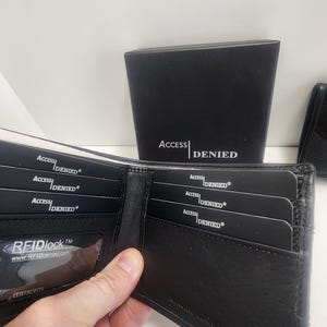 May include: Black leather wallet with multiple card slots, some containing cards with "ACCESS DENIED" printed on them. The wallet is open, revealing the interior. A black box with "ACCESS DENIED" is in the background.