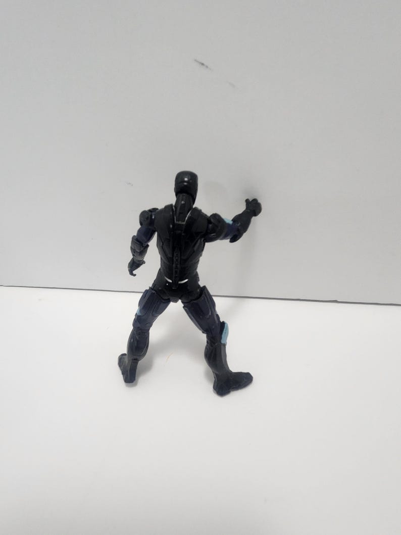 May include: Black action figure with blue accents, standing on a white surface. The figure is posed with one arm raised and the other arm at its side.
