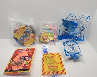 May include: A collection of six Happy Meal toys from McDonald's. The toys include a blue Smurfs character, a red and orange "Kids Choice Awards" toy, a blue and yellow "Spytracker" toy, a red and yellow "Behind the Scenes" activity book, a red and yellow "Behind the Scenes" activity book, and a blue and yellow "Behind the Scenes" activity book.