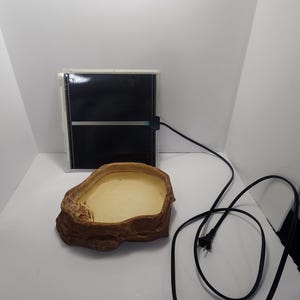 May include: A brown reptile water dish with a textured surface and a black electrical cord with a plug. The cord is connected to a black heating pad.