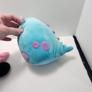 May include: A blue dinosaur-shaped plush toy with purple detailing. The toy features three large purple circles and small grey spikes. The soft toy is designed to be cuddled and is suitable for children.
