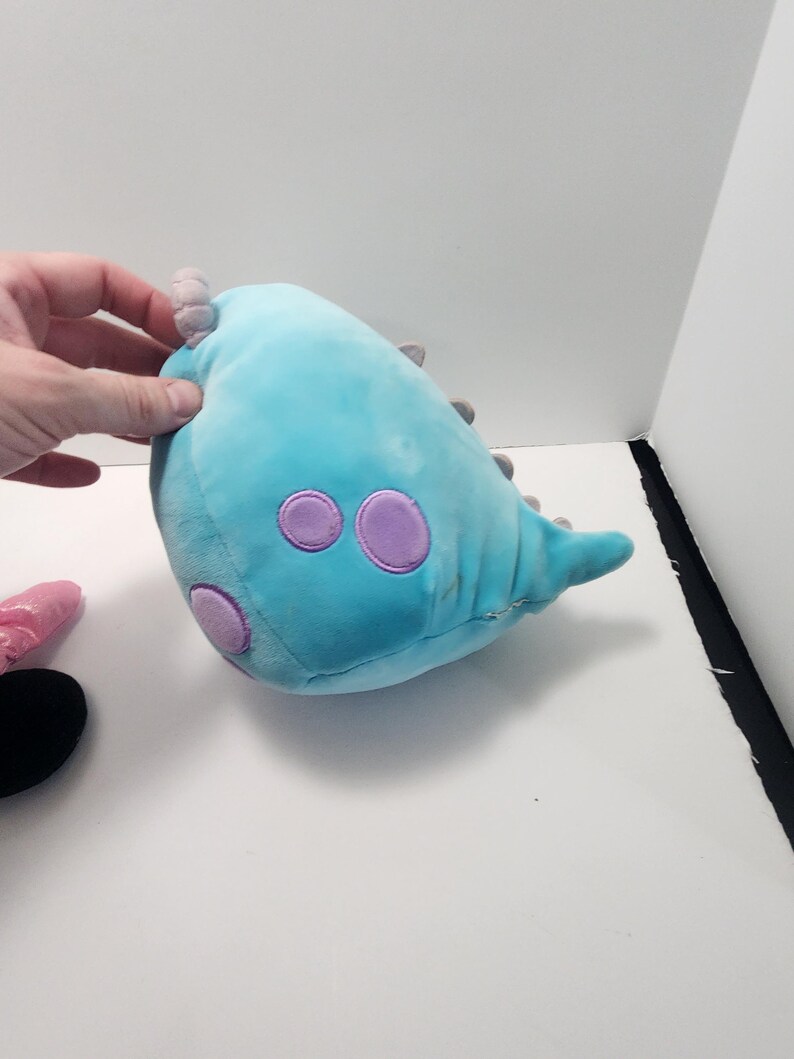 May include: A blue dinosaur-shaped plush toy with purple detailing. The toy features three large purple circles and small grey spikes. The soft toy is designed to be cuddled and is suitable for children.